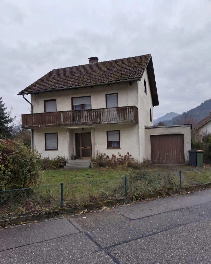 Vorher: Handyfoto eines älteren Einfamilienhauses aus den 1970er-Jahren mit Holzbalkon und Garage